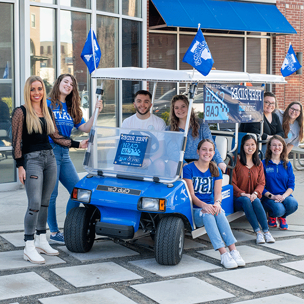 Nine Indiana State students, sit on or stand around the Cash Cab, a golf cart decorated with blue signage advertising free rides as part of Give to Blue Day. There is one male student in the image; he is seated behind the wheel of the golf cart. Several students are wearing blue Indiana State shirts.